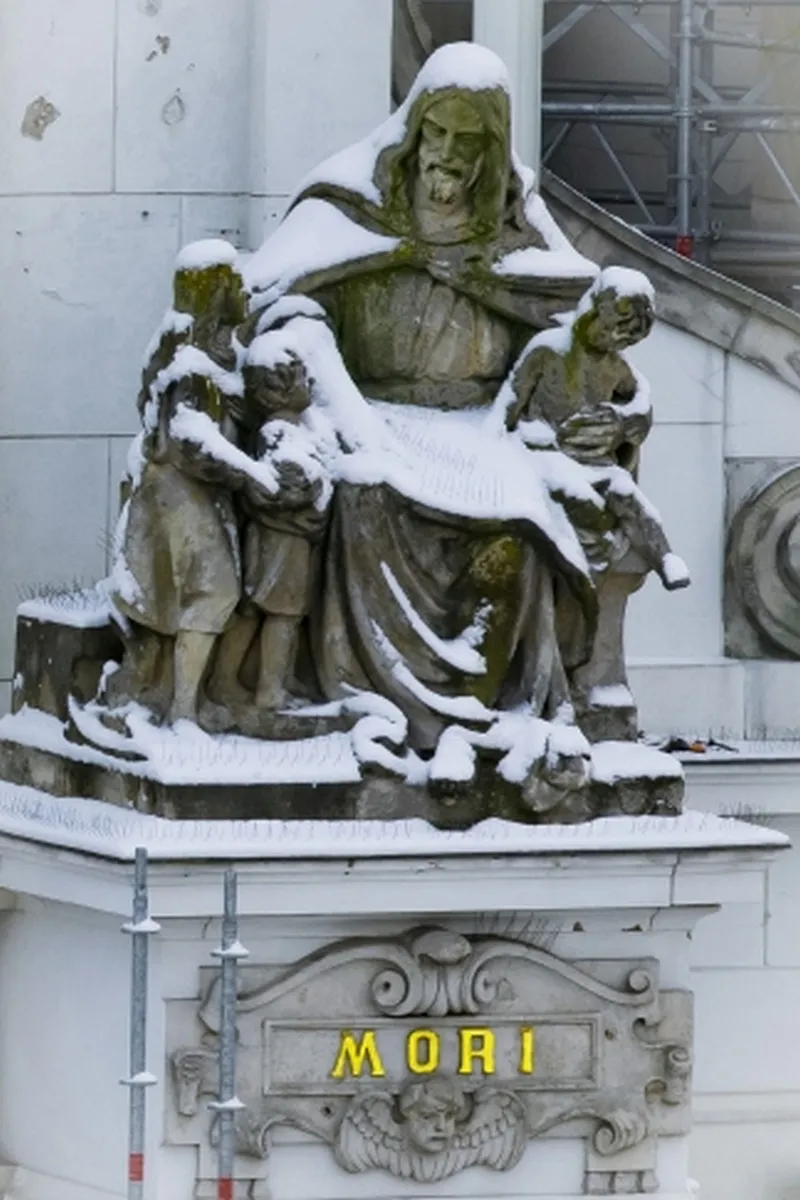 Snow-covered robed statue with children above the glowing word MORI